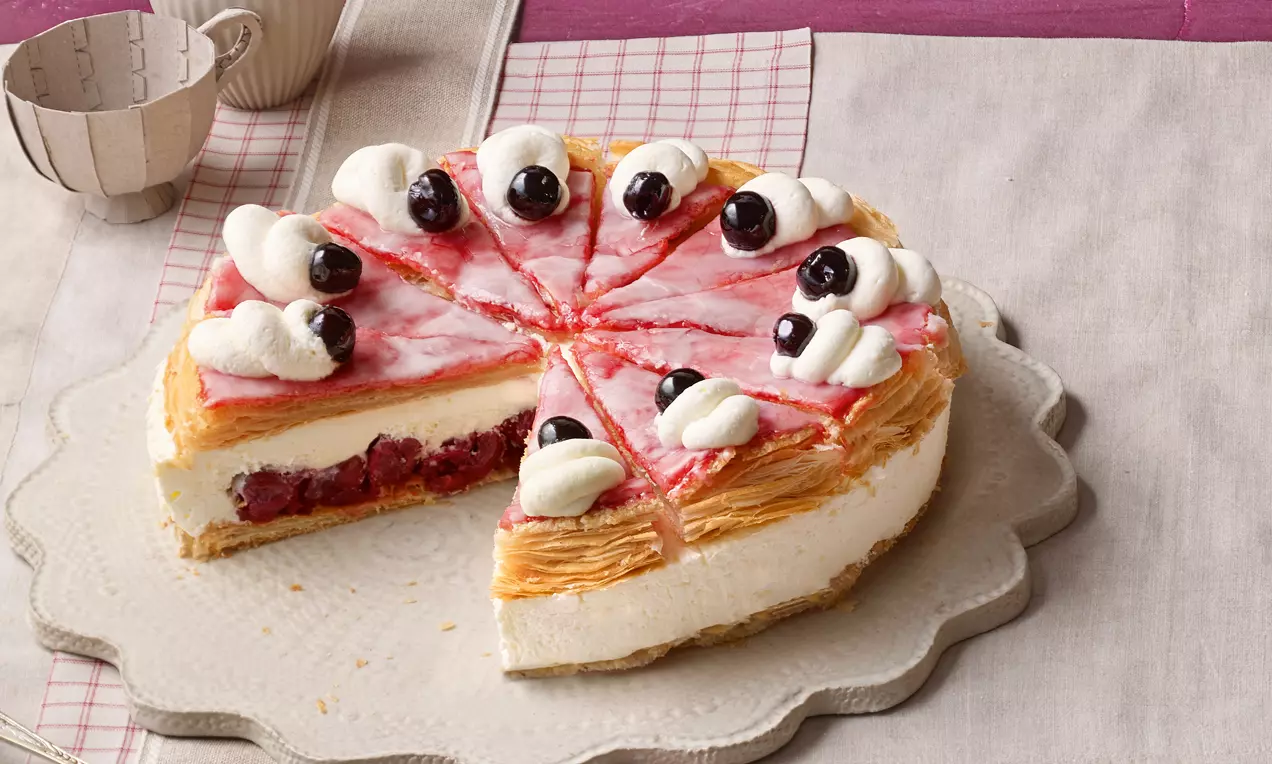 Holländer-Kirsch-Torte mit knusprigem Blätterteig, heller Creme, roter Kirschfüllung, Sahnetupfen und schwarzen Kirschen auf einer Keramikplatte