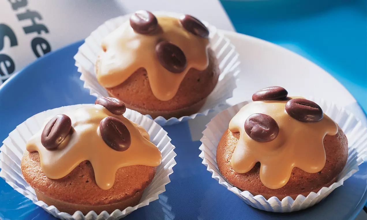 Arabica-Cupcakes mit glatter beige-goldener Glasur und schokoladenbraunen Kaffeebohnen, angerichtet in weißen Papierförmchen auf blauem Teller