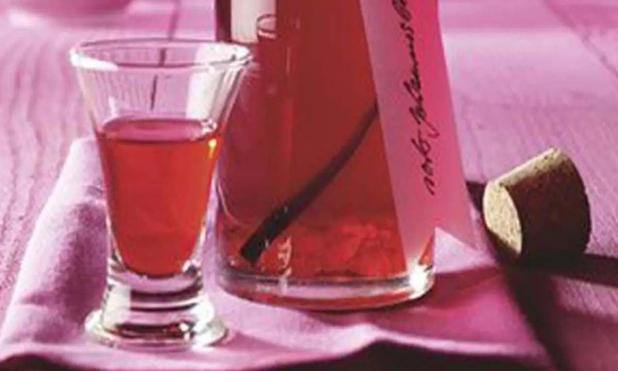 Roter Ribisellikör in einer klaren Flasche mit handgeschriebenem Etikett und Korken, daneben ein Glas auf lilafarbenem Stoff drapiert