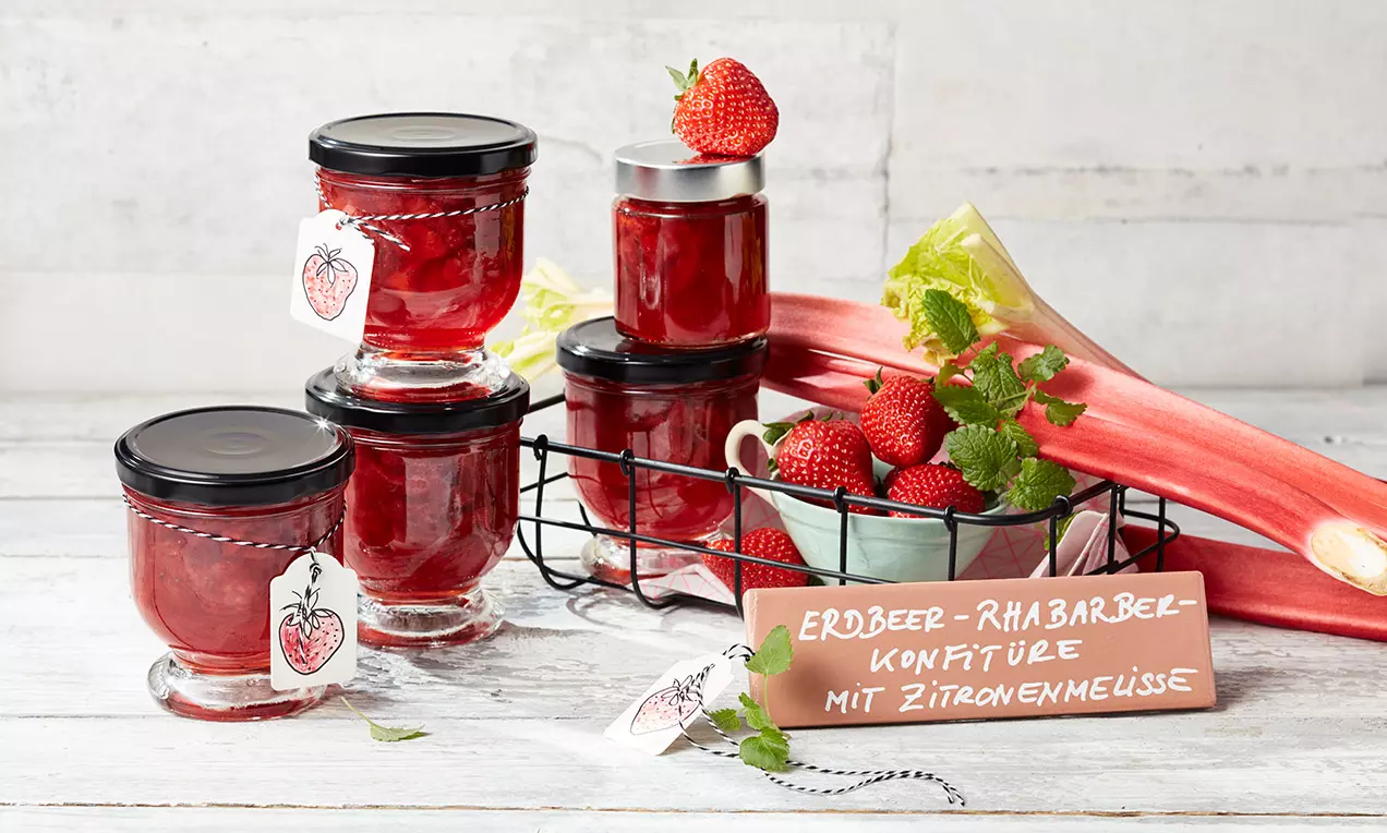 Erdbeer-Rhabarber-Konfitüre in gläsernen Schraubgläsern mit schwarzen Deckeln, dekoriert mit frischen Erdbeeren und Zitronenmelisse in Drahtkorb