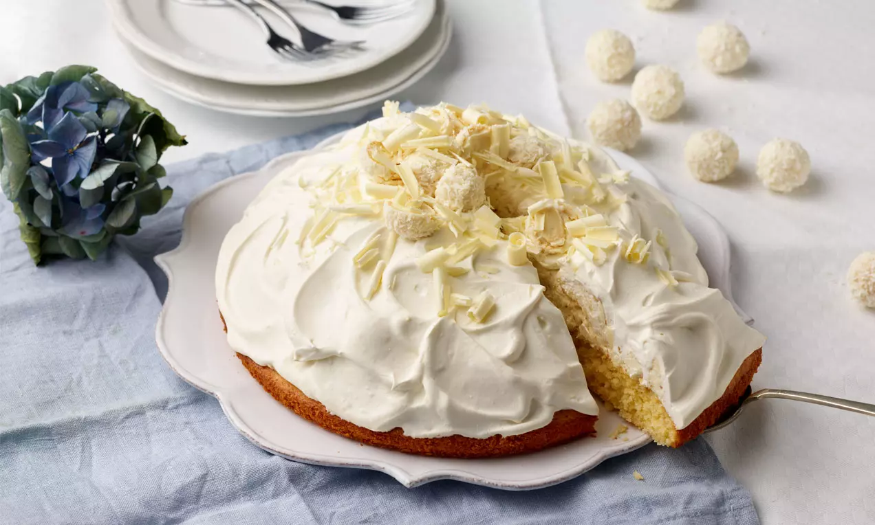 Raffaelo-Torte mit weißer Sahnecreme, bestreut mit weißen Schokoraspeln und Raffaelo-Kugeln, serviert auf einem weißen Teller mit angeschnittener Ecke