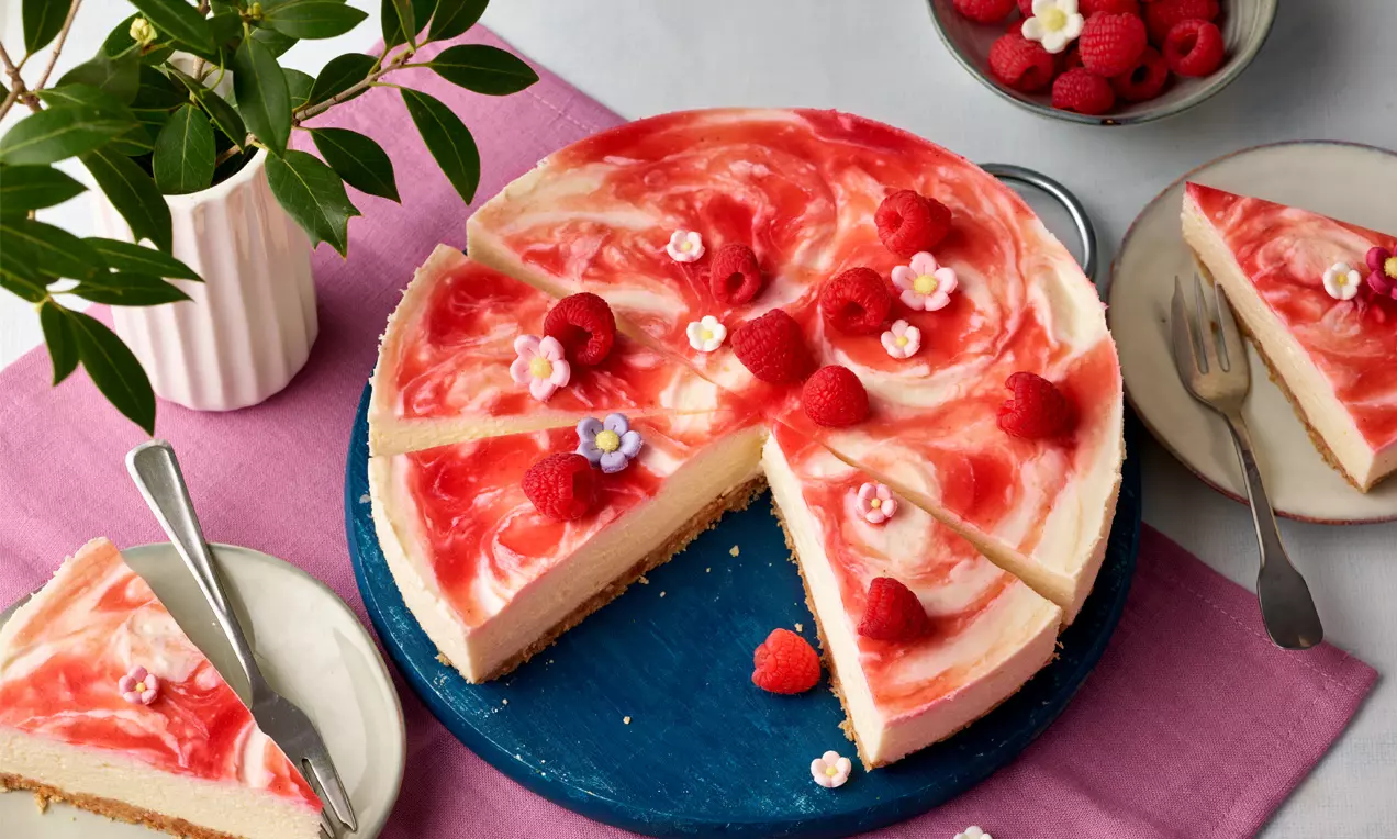 Himbeer-Käsekuchen auf blauem Teller, mit rotem Himbeerspiegel, frischen Himbeeren und Blüten dekoriert, angeschnitten auf hellem Tuch