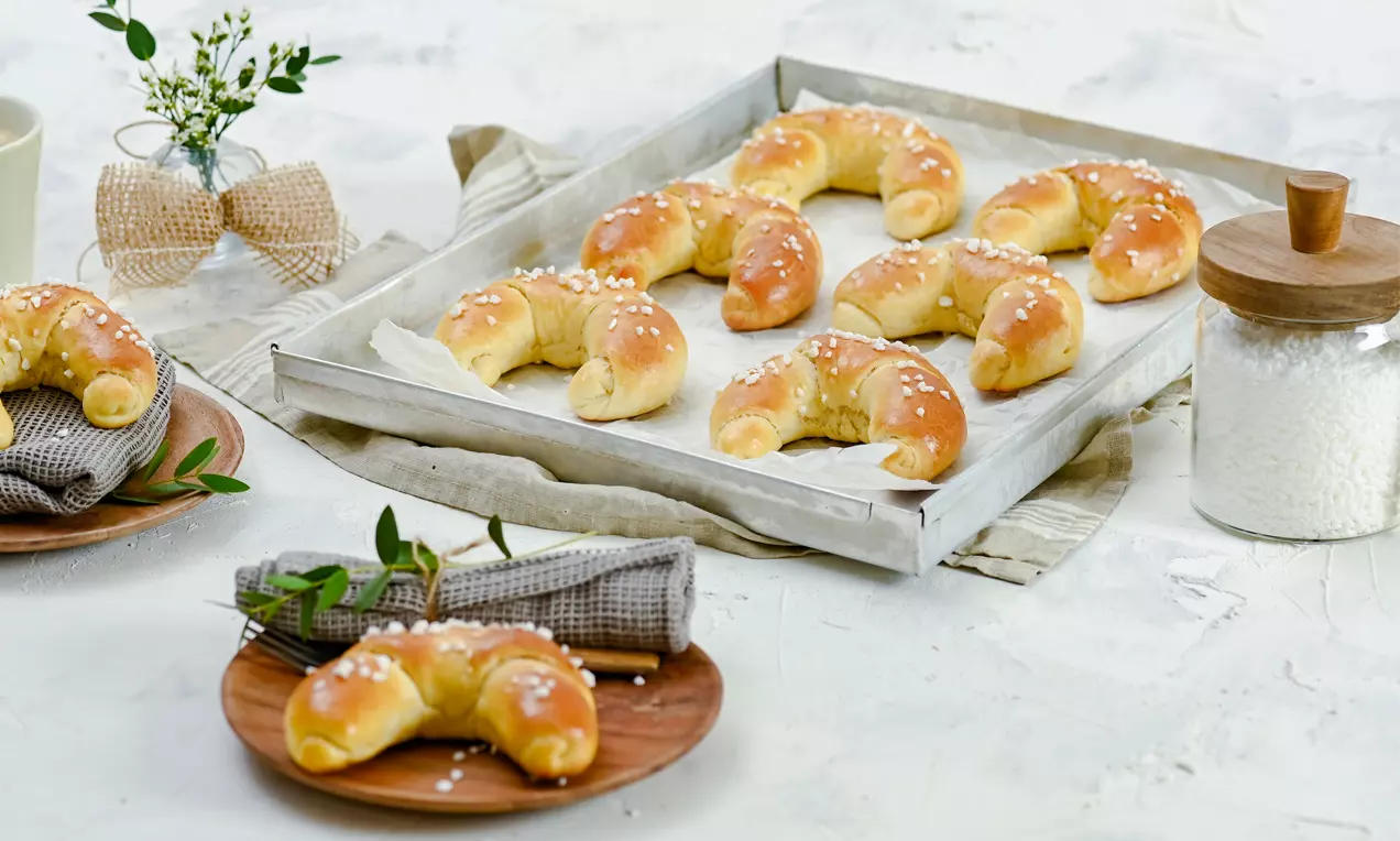 Goldbraune Frühstückskipferln mit glänzender Oberfläche und grobem Zucker bestreut auf einem Backblech sowie Holztellern mit Serviette drapiert