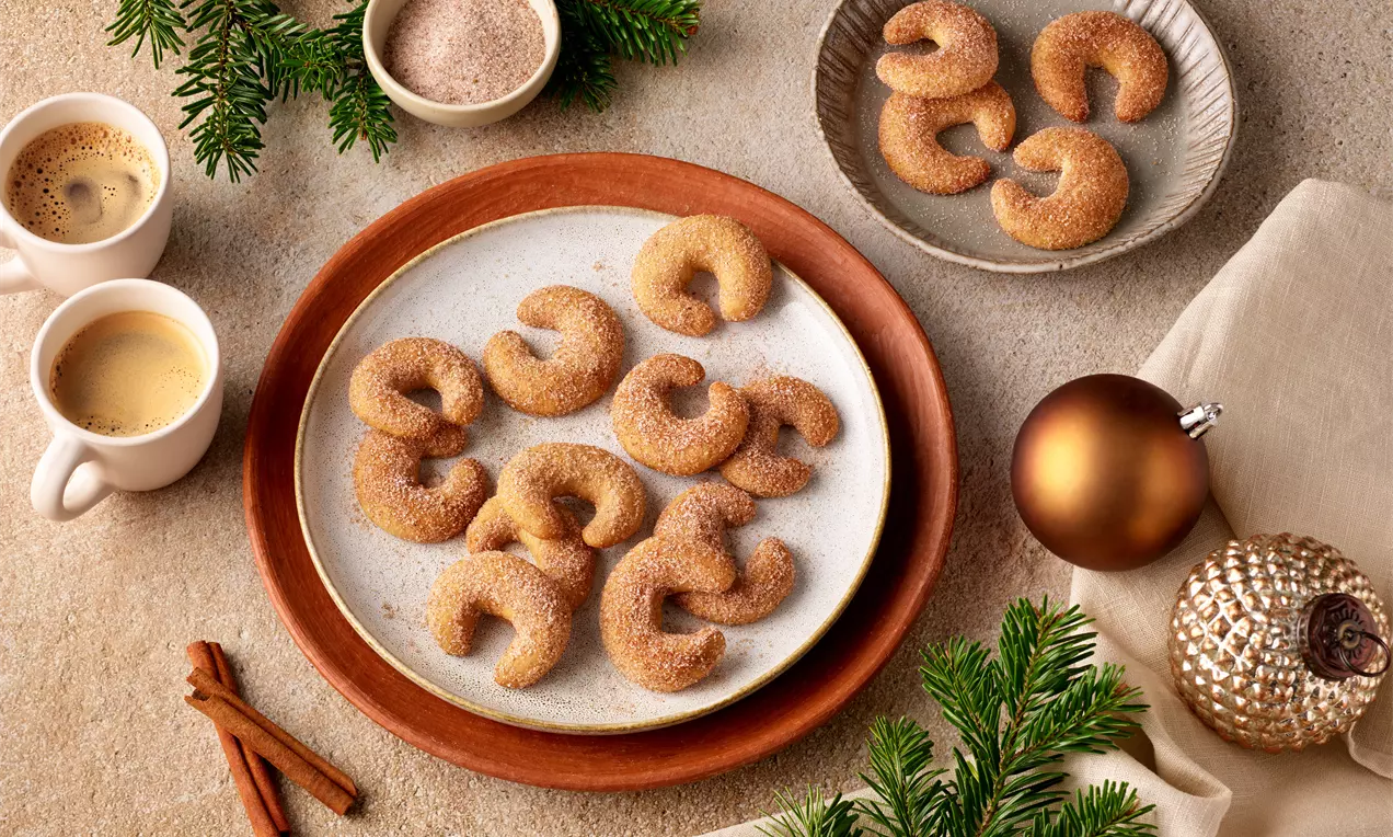 Espresso-Zimt-Kipferl, goldbraun und zu Hörnchen geformt, mit Zimt-Zucker bestreut auf Tellern zwischen Tannenzweigen und Weihnachtsdeko arrangiert