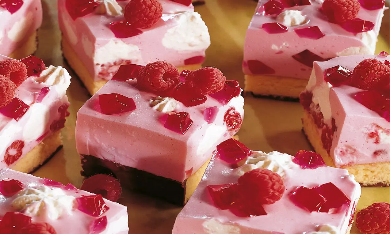 Würfel mit rosafarbener Creme und rotem Götterspeise-Gelee auf hellem Biskuitboden, garniert mit frischen Himbeeren und Sahnetupfern