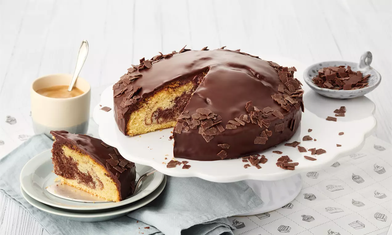 Marmorkuchen mit glänzender Schokoladenglasur und dunklen Schokoraspeln auf weißer Tortenplatte, daneben ein angeschnittenes Stück auf Teller