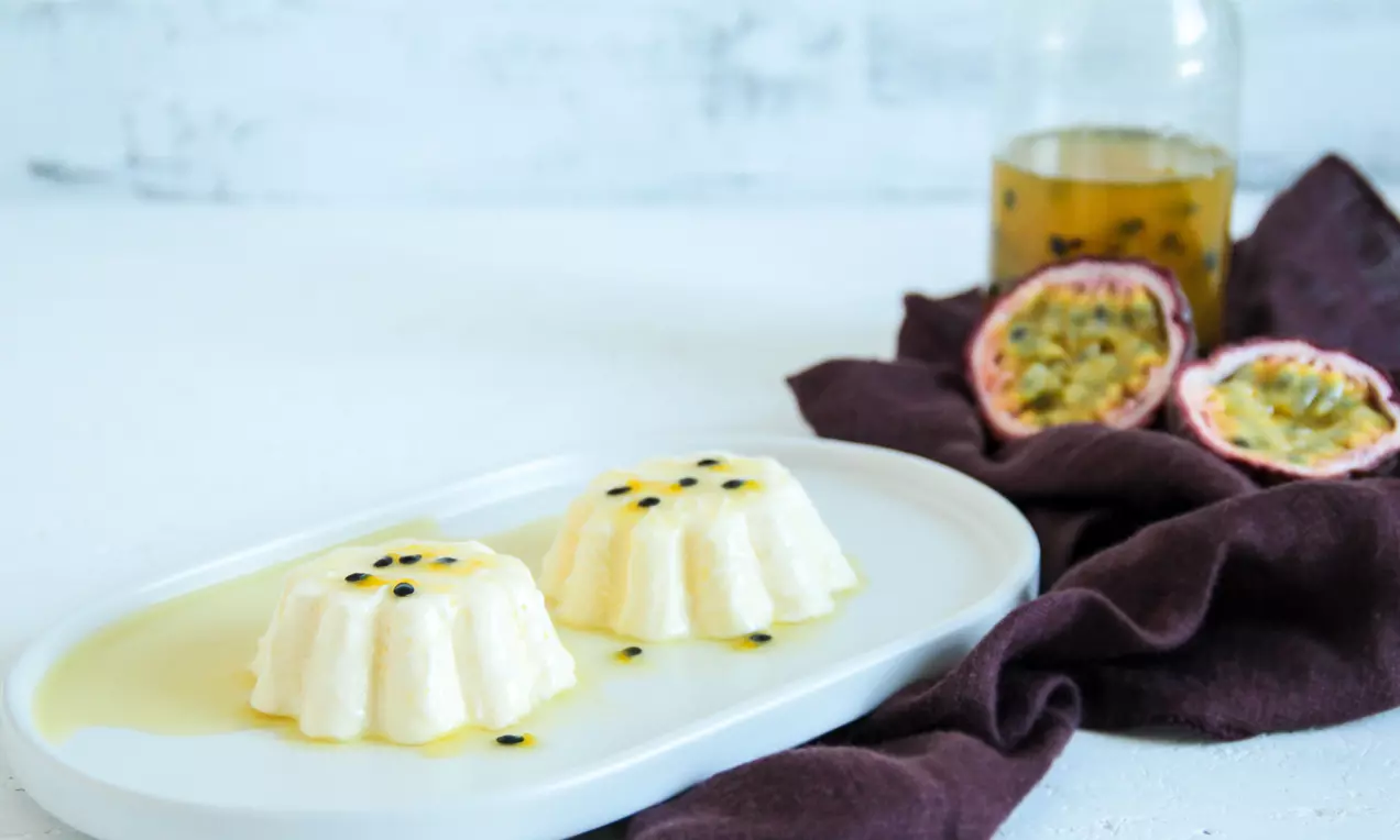 Zwei weiße, cremefarbene Schokoparfaits mit geschwungenem Rand, garniert mit Passionsfruchtkernen, auf ovaler Platte mit dunklem Stoff drapiert
