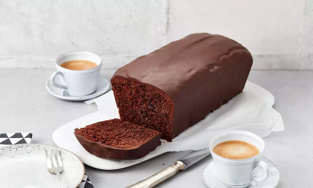 Schokoladenkuchen in Kastenform mit glänzender Schokoglasur, angeschnitten auf weißer Platte, daneben zwei Tassen Kaffee und Besteck