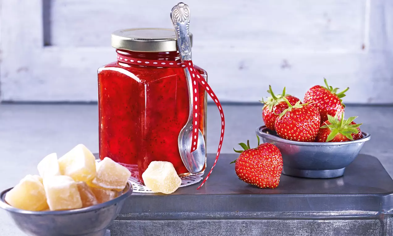 Ein Glas mit leuchtend roter Erdbeer-Ingwer-Konfitüre mit rotem Band und silbernem Löffel, daneben frische Erdbeeren und kandierter Ingwer in Schalen