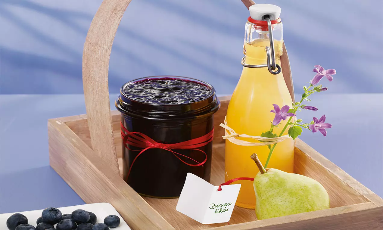 Glasflasche mit gelbem Birnenlikör neben grün-gelber Birne und lila Blüten in einem hellen Holzkorb auf blauem Hintergrund