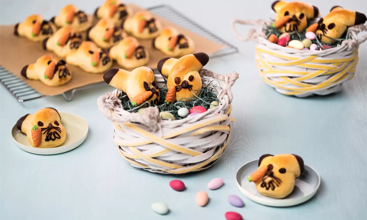 Goldbraune Gebäck-Häschen mit Schoko-Ohren und Gesicht, teils in Körbchen mit Ostergras und bunten Eiern, teils auf Tellern oder Gitterablage