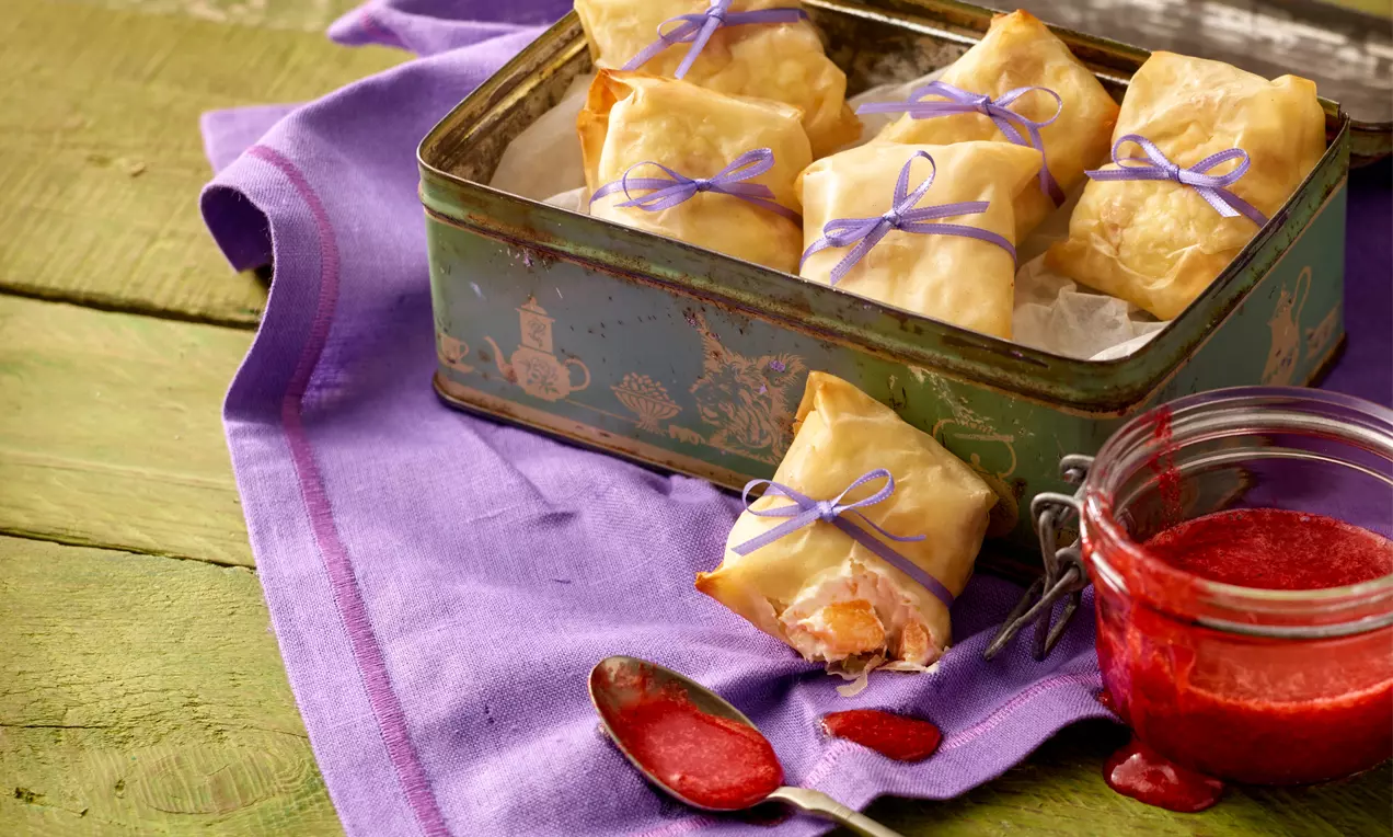 Knusprige, goldbraune Strudelteig-Päckchen mit lila Bändern verziert, in einer Blechdose auf violetter Serviette mit roter Beerensoße serviert