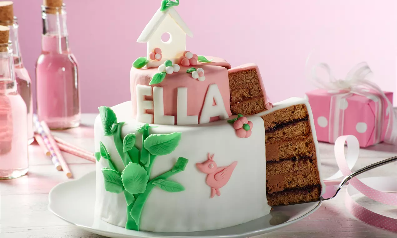 Zweistöckige Torte mit weißem Fondant, rosa Deko und Namenszug, grünen Blättern und Anschnitt der dunklen Schichten auf einer weißen Platte