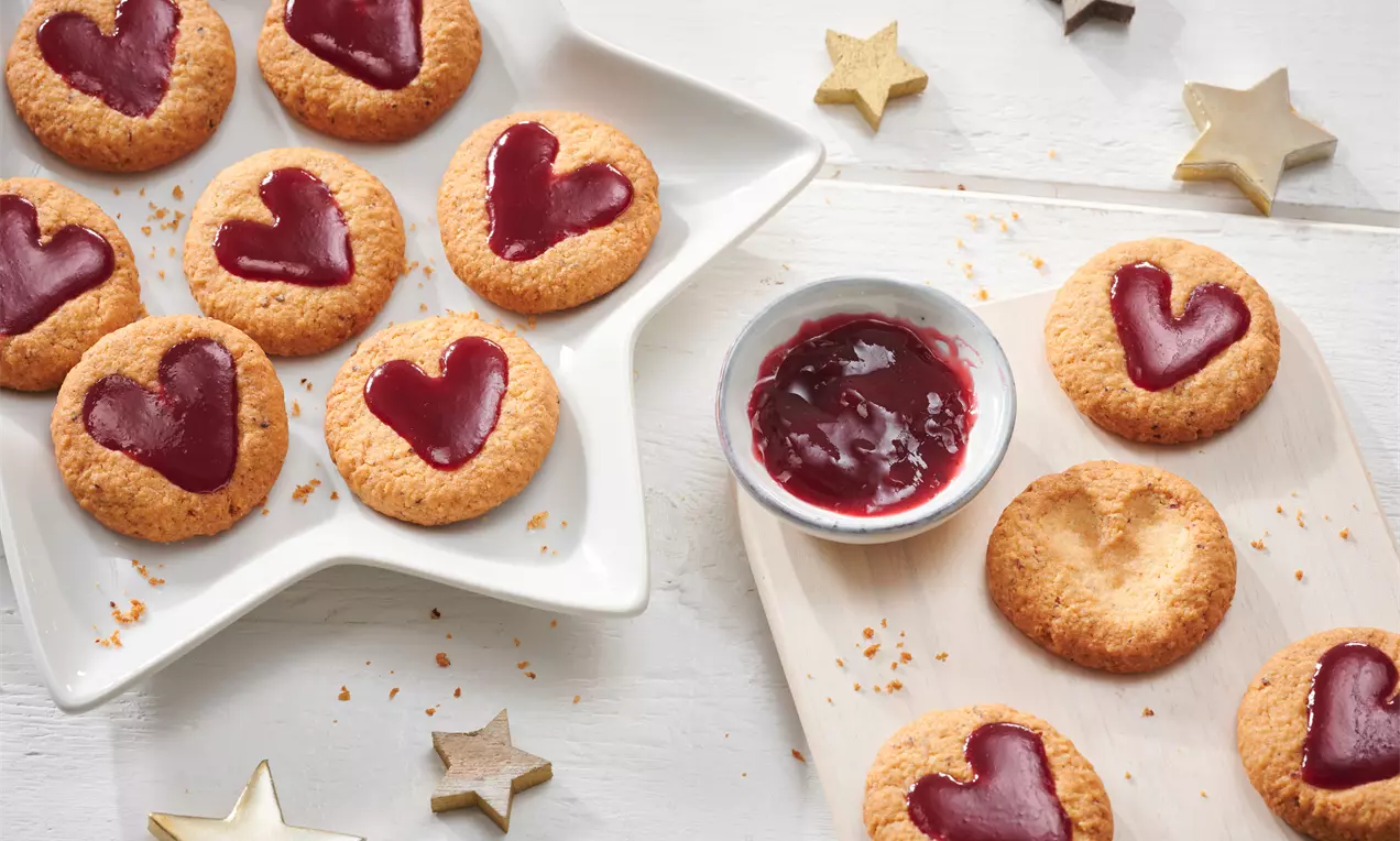 Linzer Plätzchen mit goldbrauner Kruste, herzförmiger roter Marmeladenfüllung auf einem weißen Stecksternteller und Holzbrett mit Dekosternen