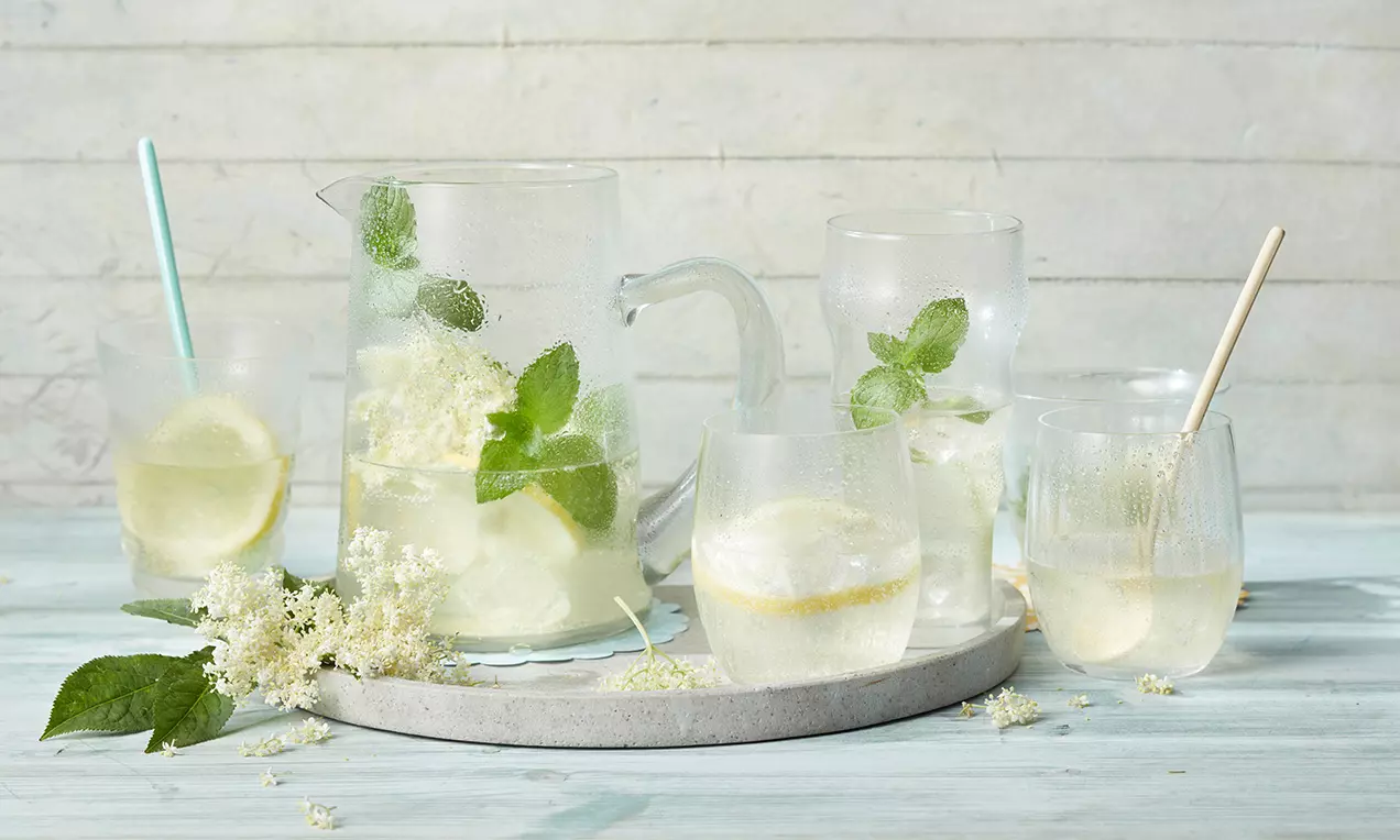 Holunderblüten-Limonade in einer gläsernen Karaffe mit Minzblättern, Zitronenscheiben und Holunderblüten, serviert in Gläsern auf hellem Tablett