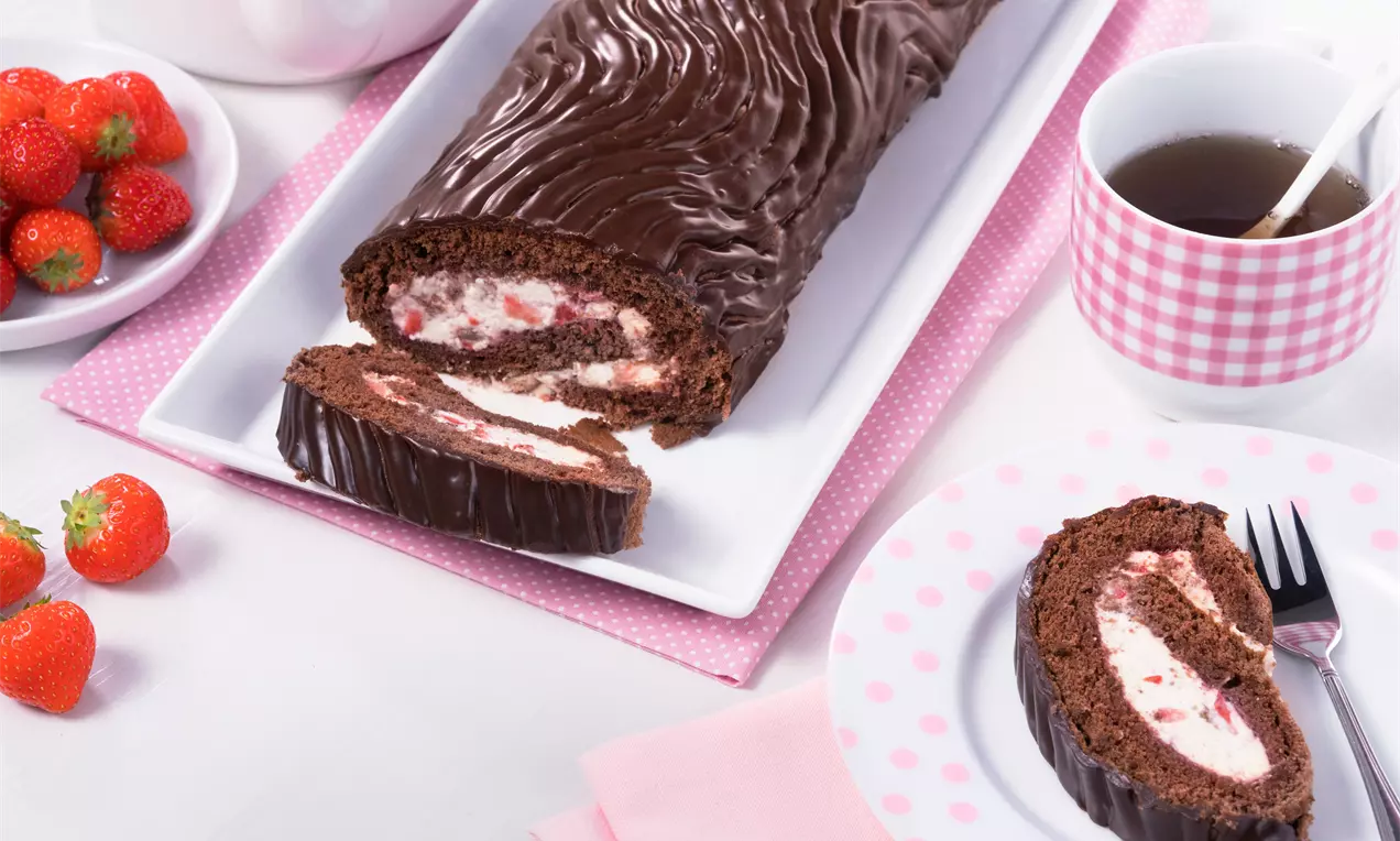 Schokoladige Biskuitrolle mit welliger Glasur, gefüllt mit weißer Creme und roten Erdbeerstückchen, auf weißem Teller angerichtet