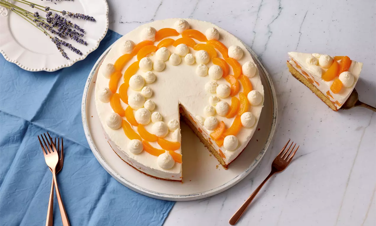 Marillen-Ricotta-Torte mit heller Creme, Sahnetupfen und Marillenstücken dekoriert auf weißem Teller neben blauem Stoff und Kupferbesteck