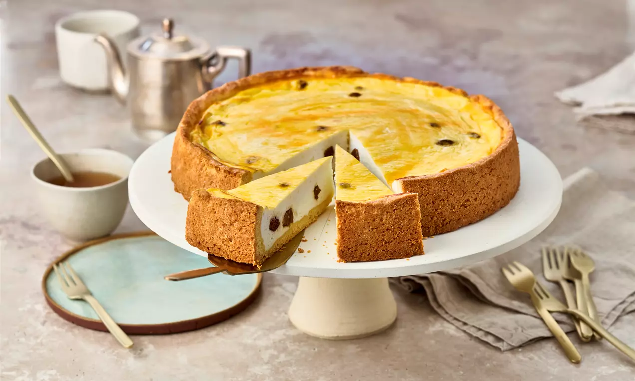 Goldgelber Käsekuchen mit Rosinen und braunem Mürbeteigrand, angeschnitten auf weißer Tortenplatte, umgeben von Geschirr und Besteck