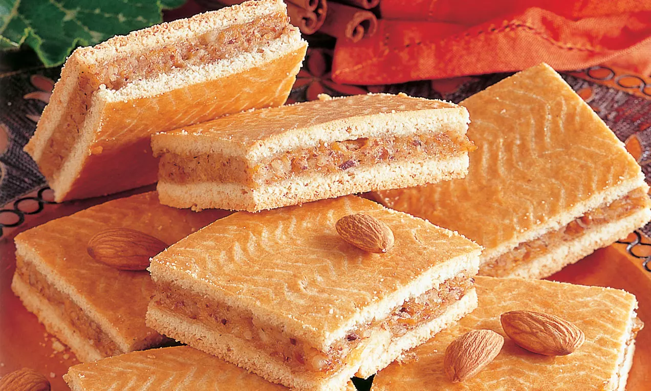 Rechteckige Kürbiskuchenstücke mit orange-goldener Oberfläche und feinem Muster, gefüllt mit braunem Kürbis und Nüssen, garniert mit Mandeln