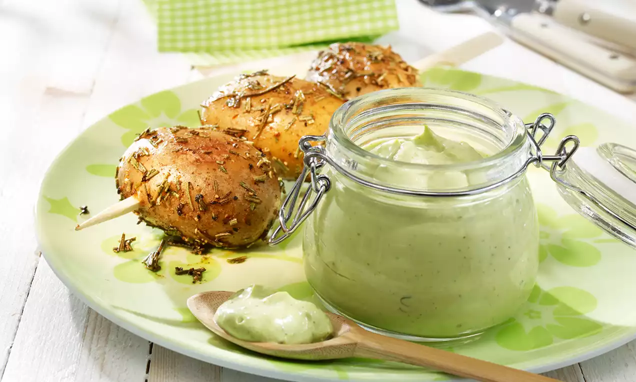 Goldbraune Kartoffelspieße mit Kräutern auf grün gemustertem Teller, daneben Glas mit cremigem grünen Avocado-Basilikum-Dip