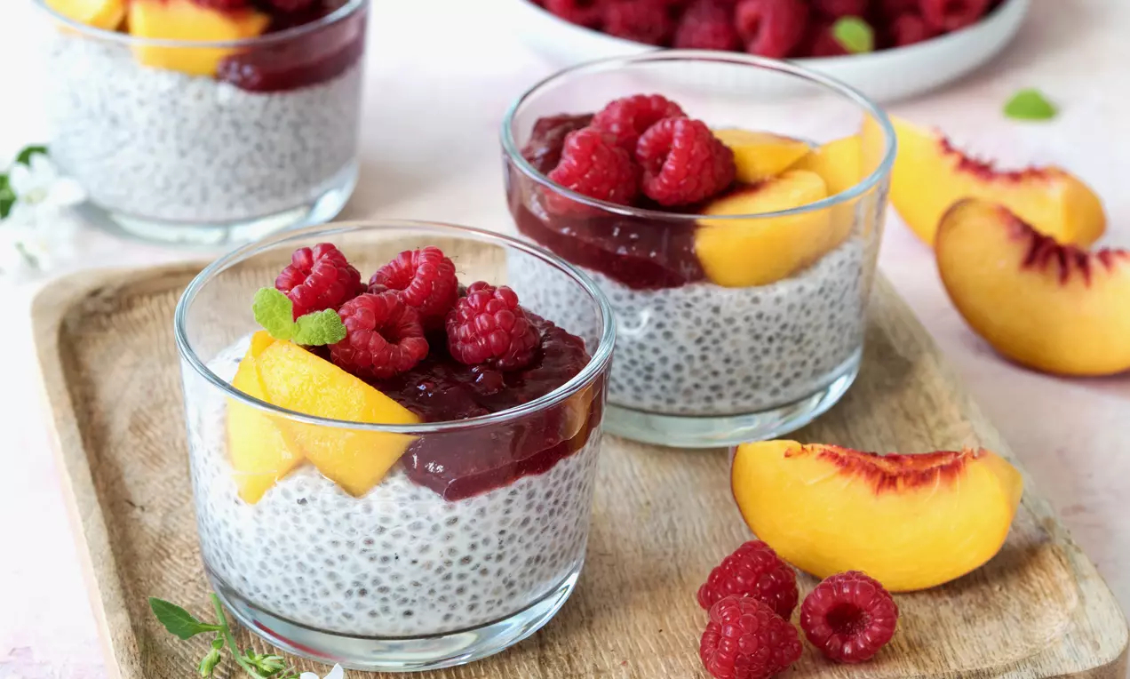 Chia-Pudding in klarem Glas, bedeckt mit dunkelrotem Fruchtmus, goldgelben Pfirsichspalten, frischen roten Himbeeren und Minzblättern