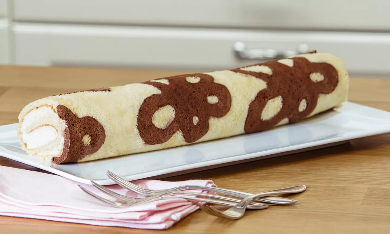 Helle Biskuitroulade mit dunkelbraunem Blümchenmuster, weißer Füllung, dekorativ angerichtet auf rechteckigem Teller mit rosa Serviette und Gabeln