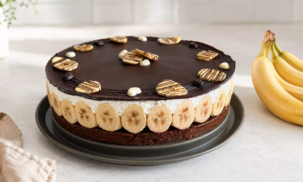 Runde Schoko-Bananen-Topfentorte mit glänzendem Schokoguss, dekoriert mit Bananenscheiben und abwechselnden Schokostreifen, AI-labeled image
