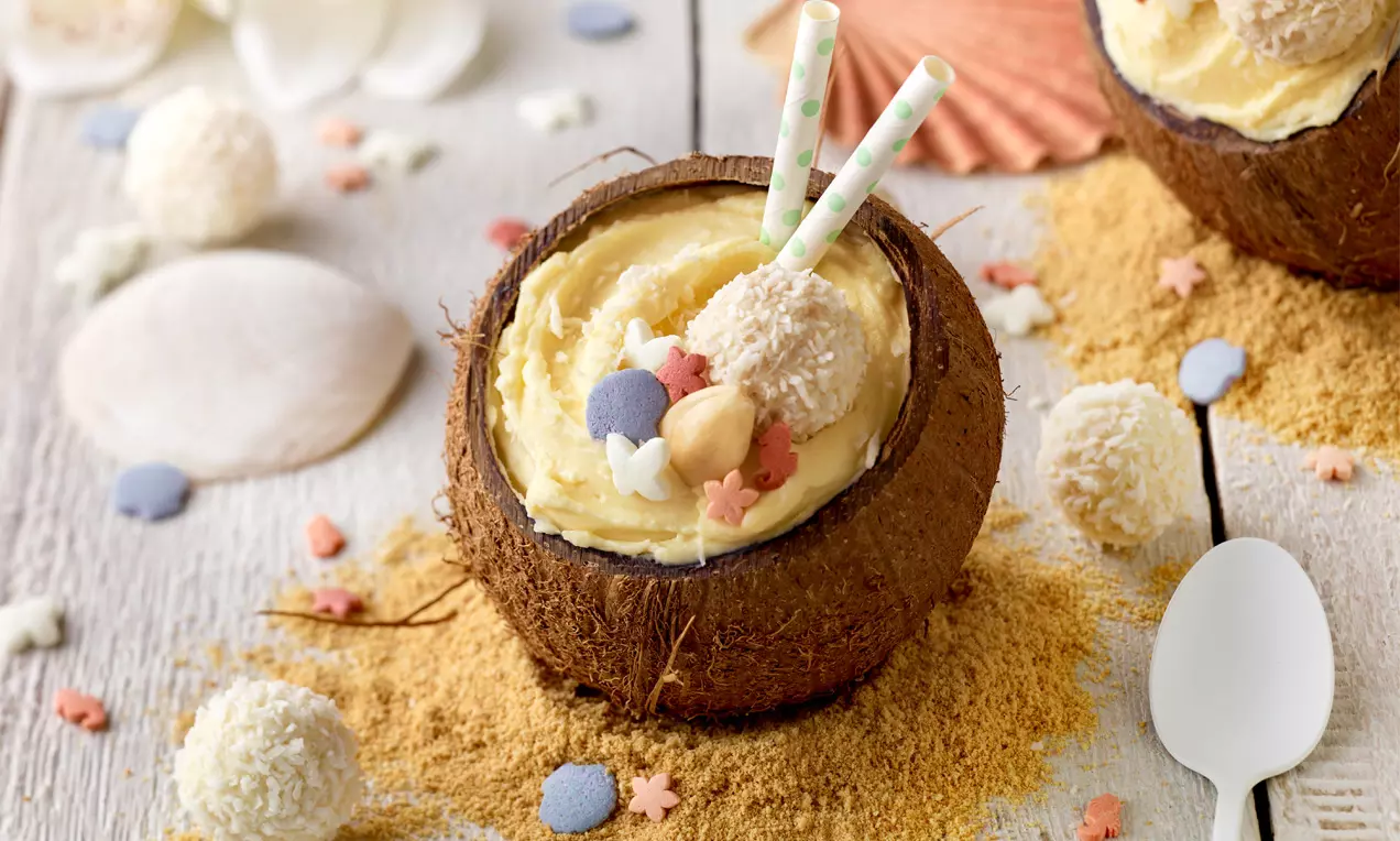 Weißes Schoko-Mousse in einer halben braunen Kokosnuss, dekoriert mit einem Raffaello, bunter Streudeko und zwei gestreiften Strohhalmen