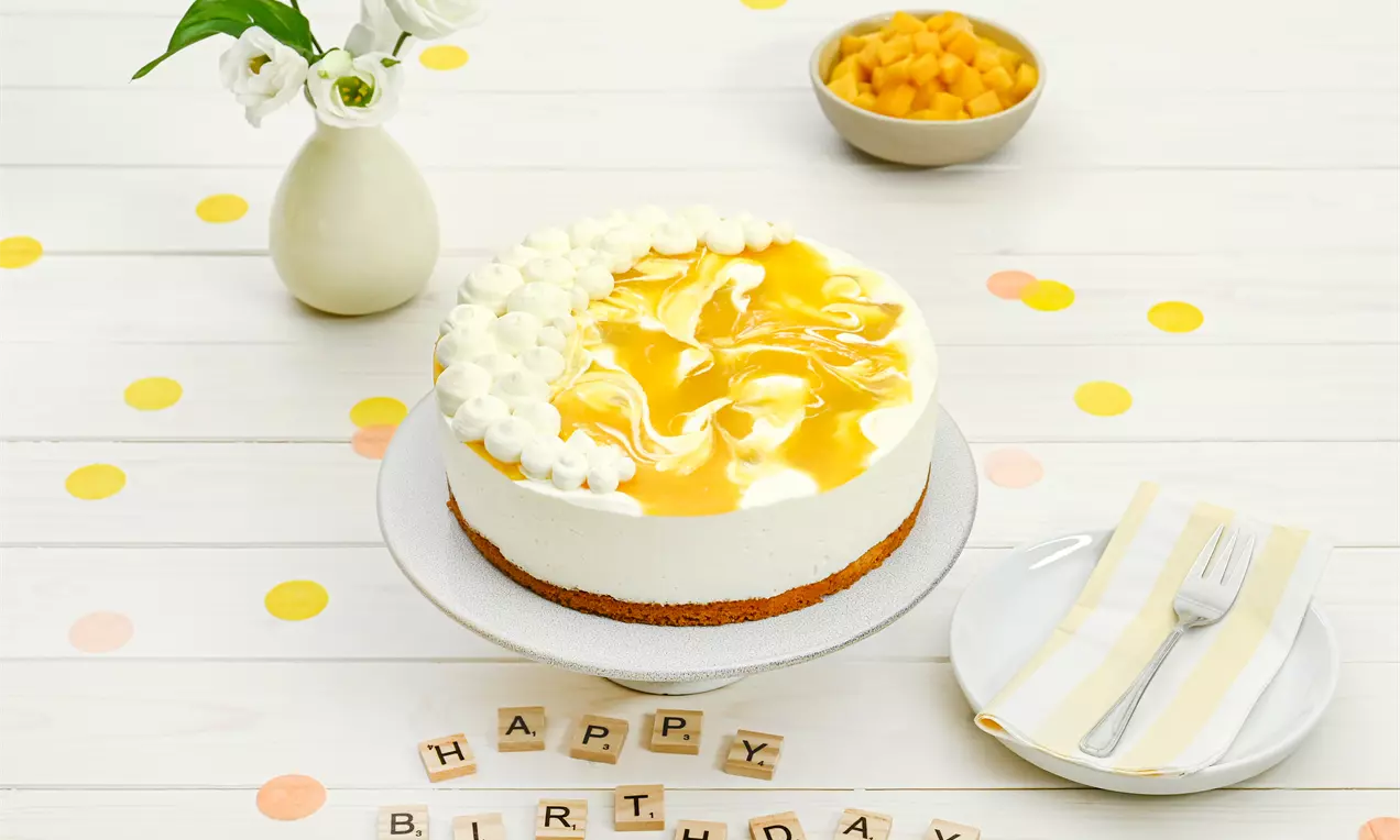 Käsesahne-Mango-Torte mit goldenem Mangospiegel, luftigem Sahnerand und braunem Boden auf einer weißen Tortenplatte aus Keramik