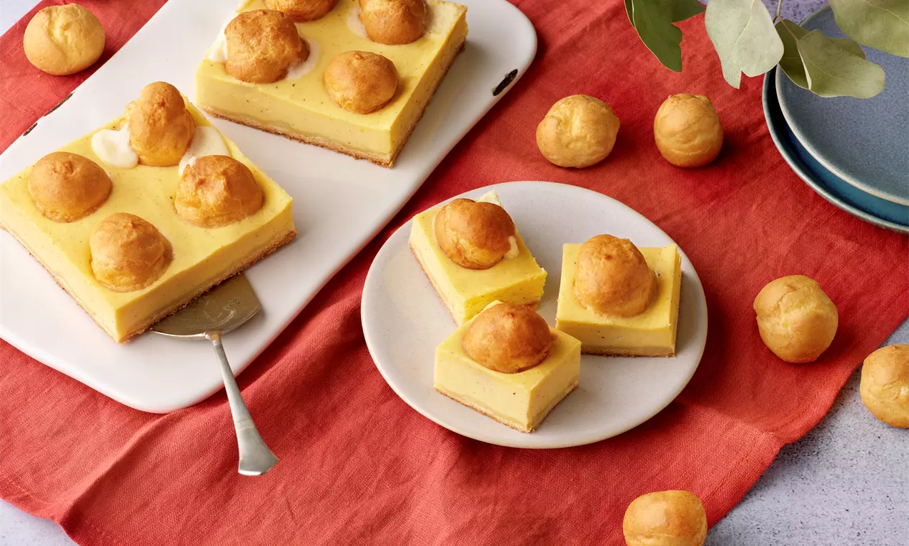 Cheesecake mit gelber Creme und goldenen Windbeuteln auf rechteckigem Blech sowie in Stücke geschnitten auf einem runden Teller