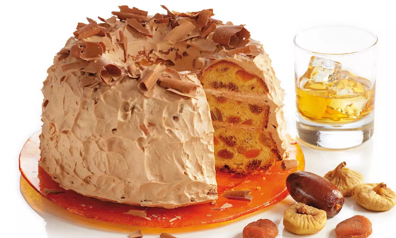 Whiskykuchen mit hellbrauner Creme, gerollten Schokospänen und einem Anschnitt, arrangiert mit Trockenfrüchten und Whiskyglas auf orangem Teller