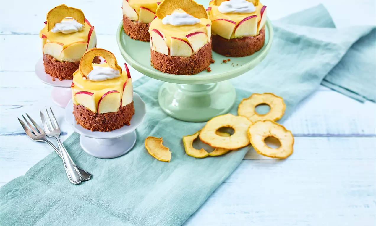 Vegane Apfeltörtchen mit goldgelber Creme auf knusprigem Boden, dekoriert mit Apfelscheiben und Sahnetupfen auf grüner Etagere drapiert