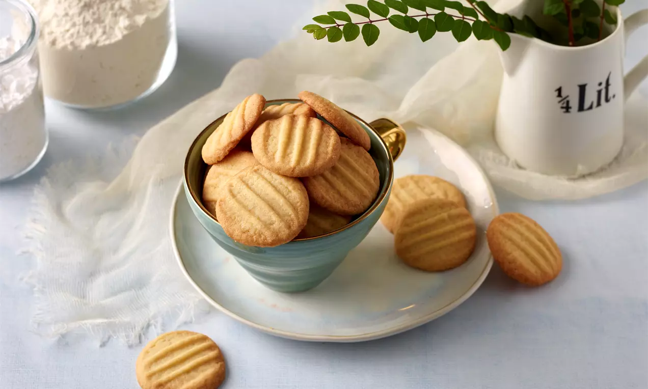 Goldbraune, runde Mürbteigkekse mit Rillenmuster in einer türkisfarbenen Tasse auf einem weißen Teller mit Deko aus Stoff und Grünzweig