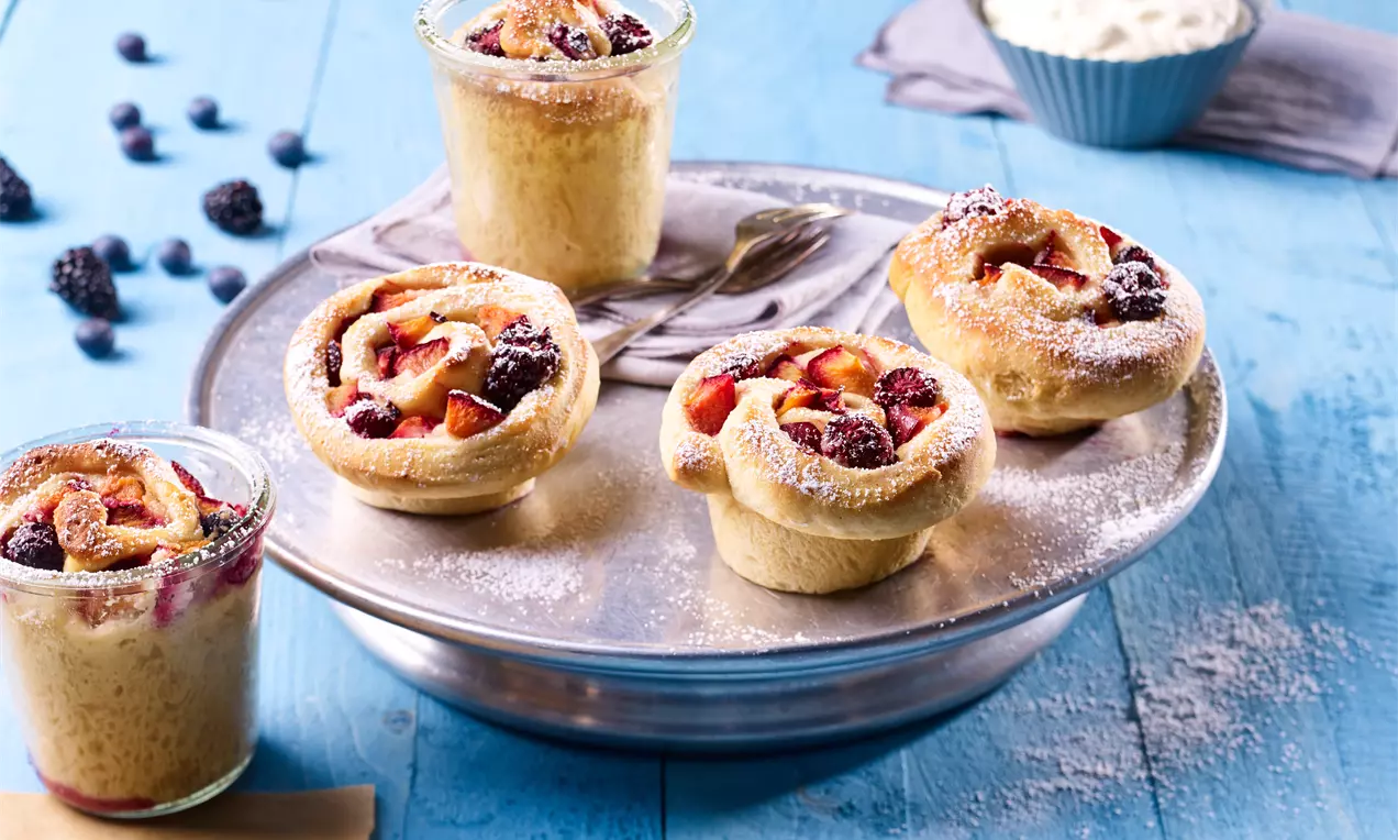 Fünf goldbraune, locker gerollte Hefeschnecken mit roten Pflaumen und dunklen Beeren bestäubt mit Puderzucker auf silbernem Teller