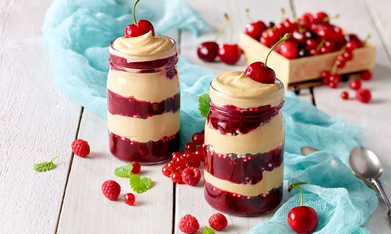 Schichtdessert aus roter Grütze und cremiger Karamellcreme in Gläsern, garniert mit Kirschen, umgeben von roten Früchten auf Holztisch