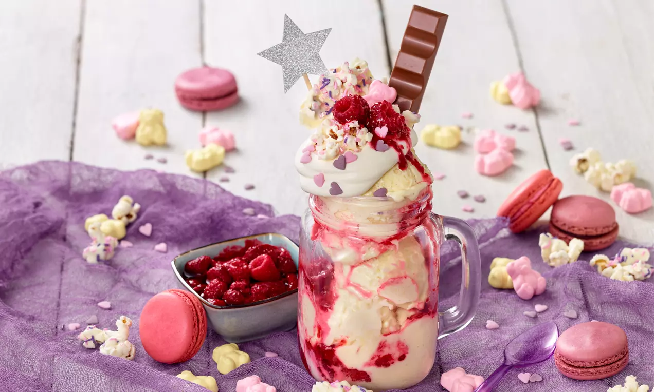 Vanilleeis mit roter Himbeersoße in einem Glas, dekoriert mit Sahne, Streuseln, Himbeeren, Schokolade und einer silbernen Stern-Deko