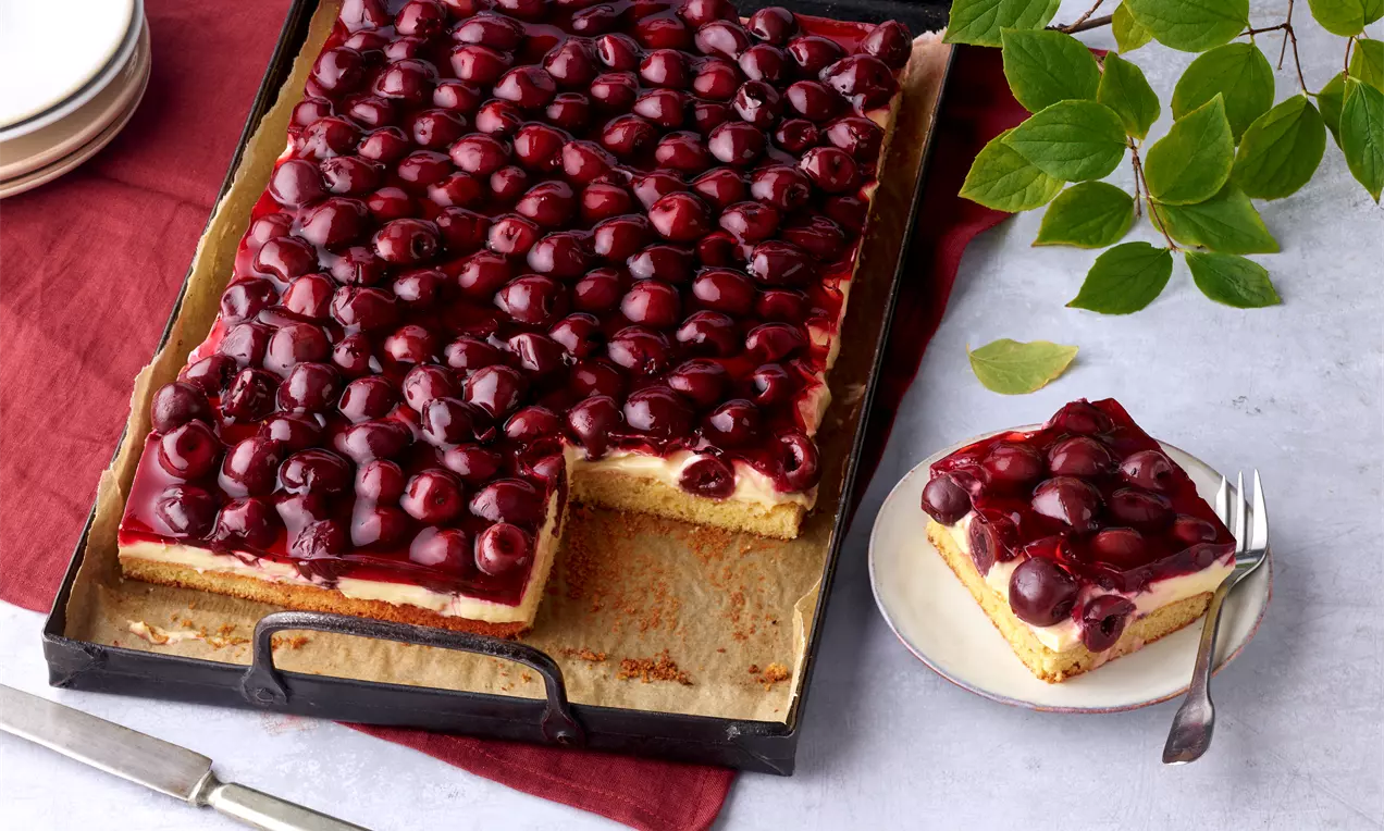 Rechteckiger Kirsch-Sauerrahm-Kuchen mit glänzend dunkelrotem Belag, auf Backpapier in einer schwarzen Form mit Stück auf weißem Teller