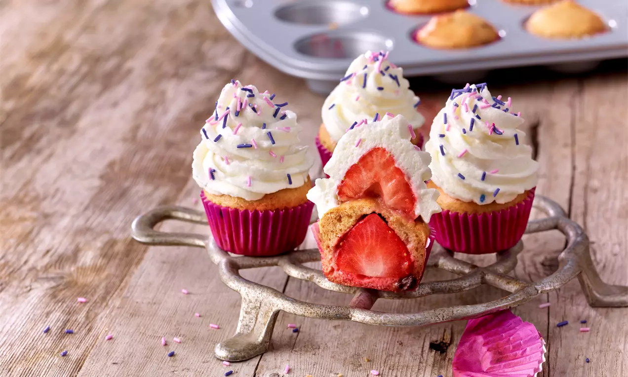 Erdbeer-Cupcakes mit Dinkelvollkornmehl, rosa Förmchen, weißem Sahnehäubchen, bunten Streuseln und frischer Erdbeere auf Metallgestell