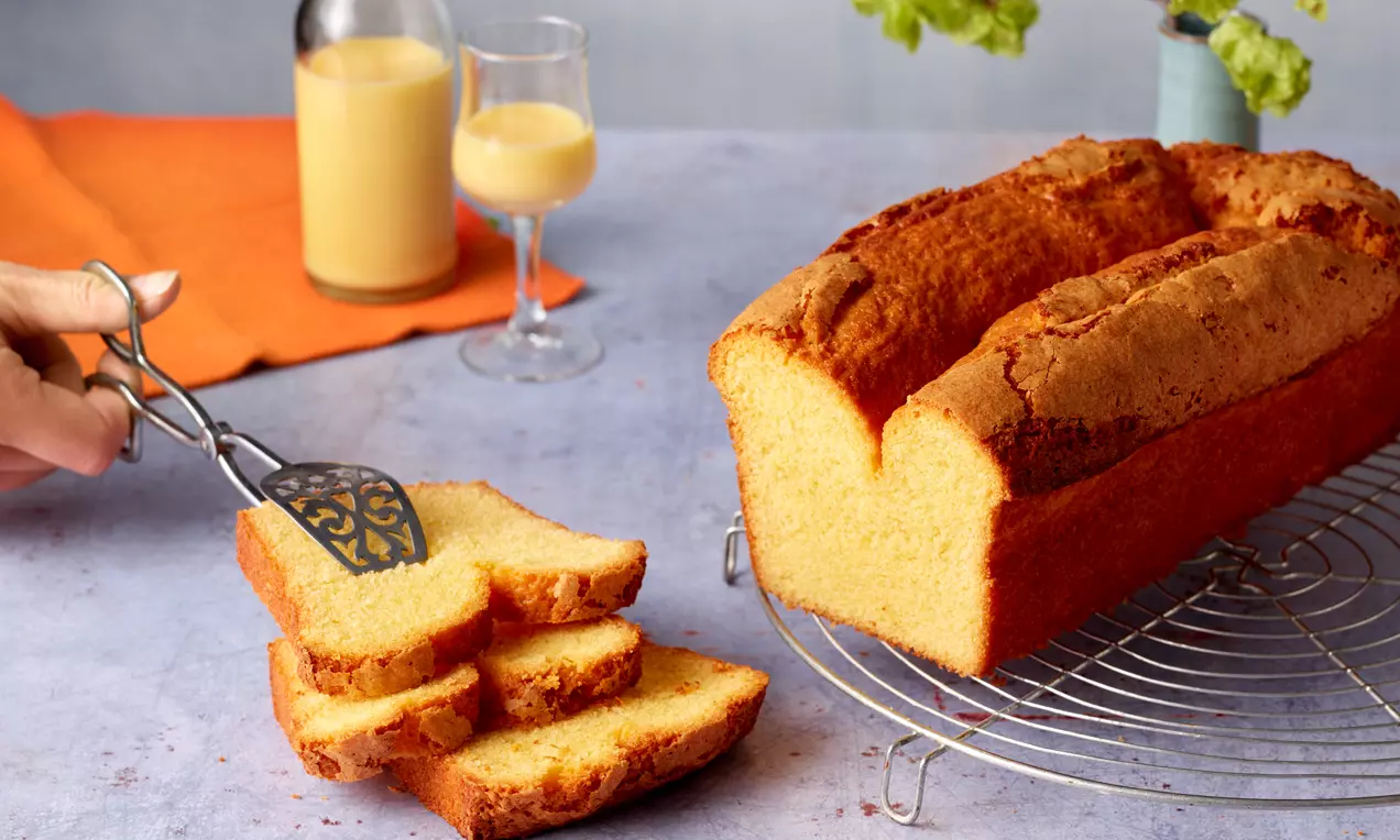Saftiger goldgelber Eierlikörkuchen mit bräunlicher Kruste auf einem silbernen Kuchengitter, daneben aufgeschnittene Stücke und Eierlikör im Glas