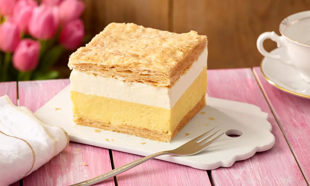 Cremeschnitten mit goldbraunem Blätterteigdeckel, heller Sahnecreme und gelber Puddingfüllung auf weißem Teller mit Gabel und rosa Untergrund
