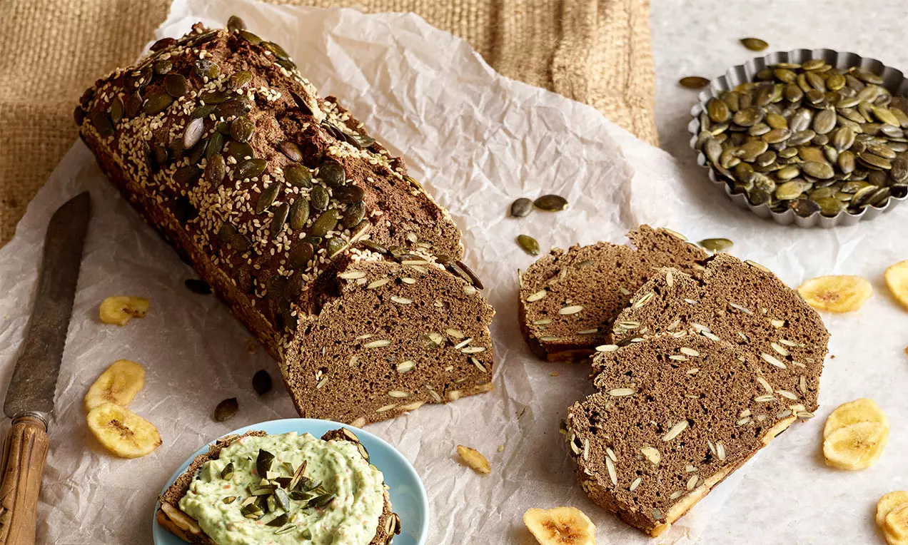 Dunkles Eiweißbrot mit knuspriger Kruste, bestreut mit Kürbiskernen und Sesam, teils in Scheiben geschnitten, auf rustikalem Pergamentpapier