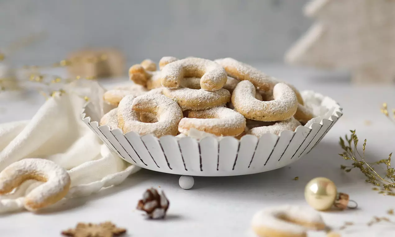 Vanillekipferl goldbraun und halbmondförmig, leicht bestäubt mit weißem Puderzucker in einer weißen Schale, umgeben von winterlicher Dekoration
