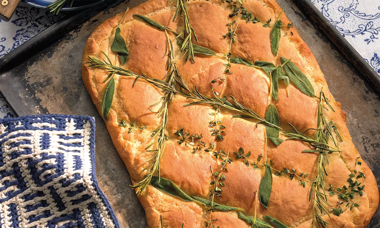 Flaches, goldbraunes Käsebrot mit eingebackenen grünen Fenchelzweigen und Kräutern auf einem dunklen Backblech neben blau-weißem Geschirrtuch