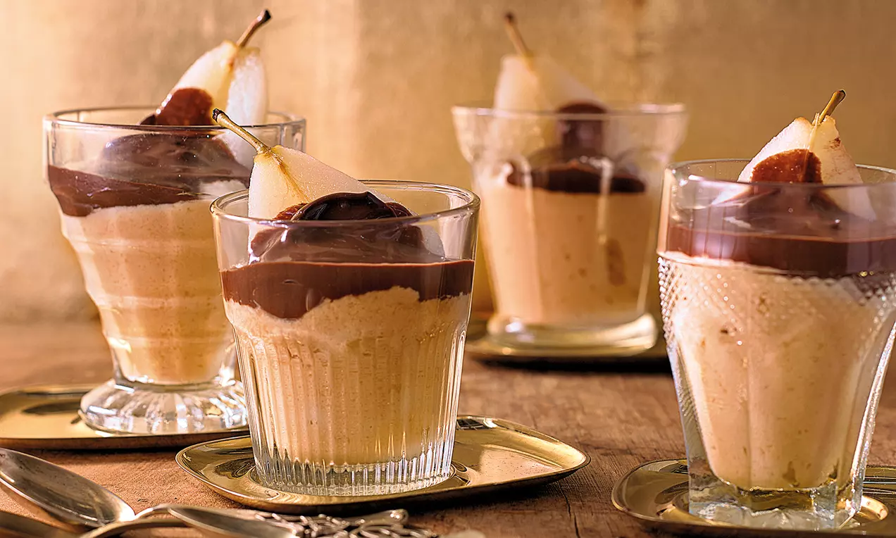 Karamellfarbenes Mousse in gläsernen Dessertschalen mit dunkler Schokosoße und hellen Birnenhälften auf goldenen Tellern angerichtet