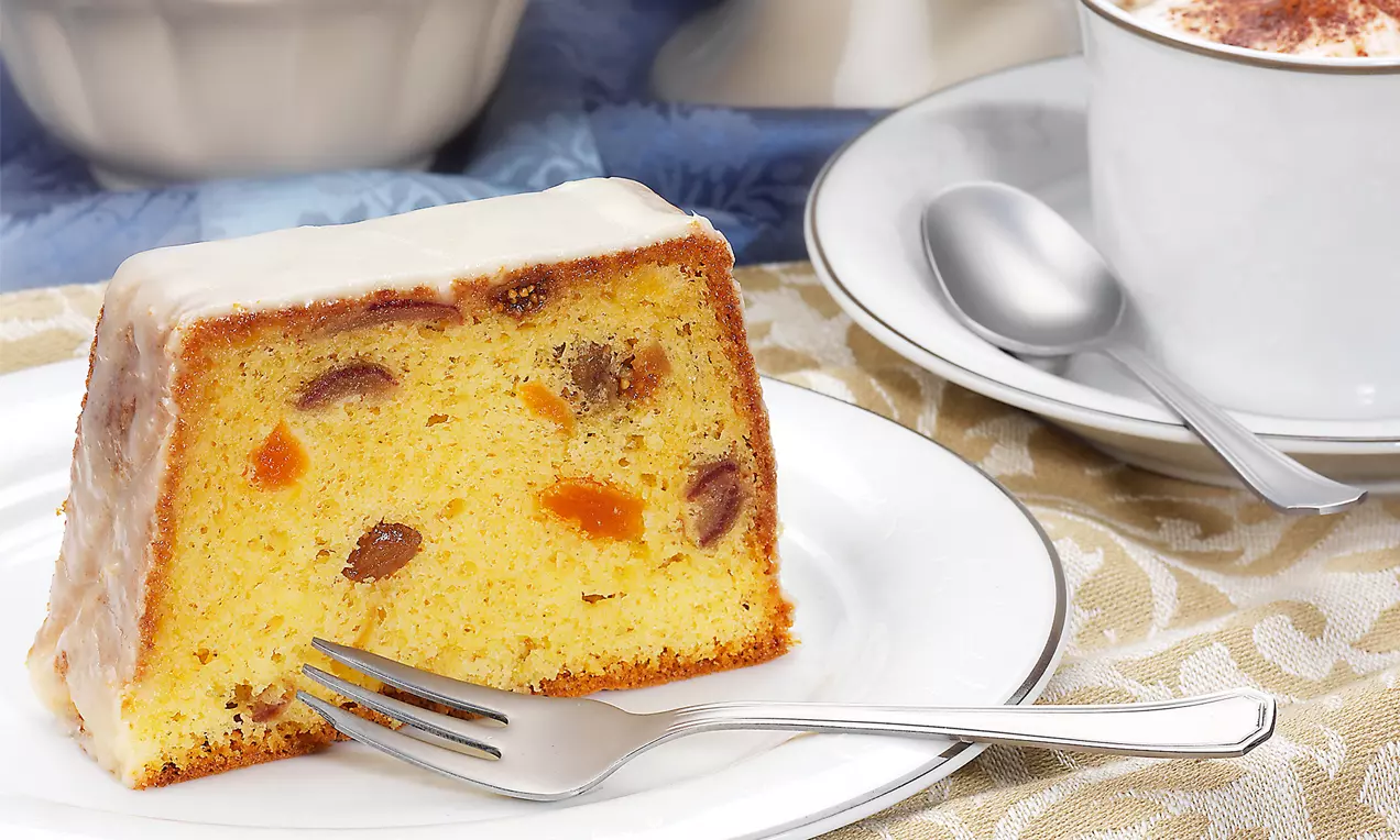 Ein Stück Königskuchen mit hellgelbem, saftigem Teig und kandierten Früchten, glatter Zuckerglasur, serviert auf einem weißen Teller mit Gabel
