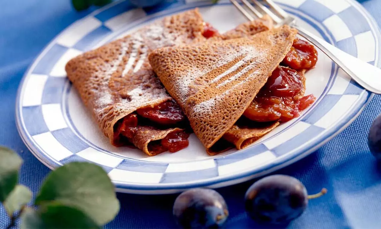 Dünne, braune Schoko-Palatschinken gefüllt mit glänzenden, rot-violetten Zwetschken, bestreut mit weißem Staubzucker auf einem Teller