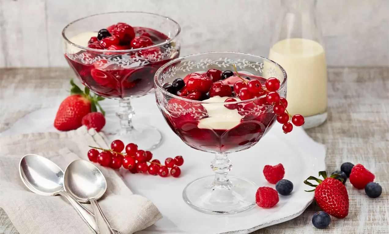 Rote Grütze aus glänzenden roten Beeren mit heller Vanillesoße serviert in verzierten Gläsern auf weißem Teller mit frischen Beeren