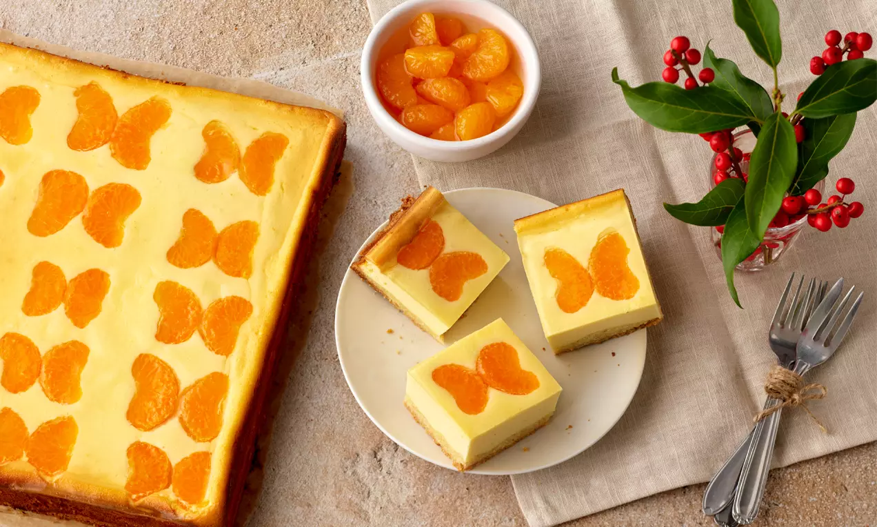 Quadratische Kuchenstücke mit gelber Creme und Mandarinen-Schmetterlingen auf hellem Teller, daneben Servietten, Besteck und Dekoration