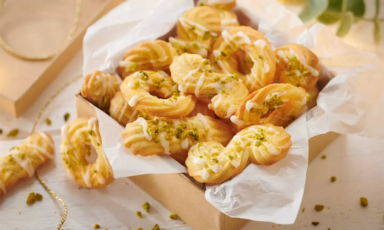 Goldgelbe, spritzförmige Orangenplätzchen mit weißem Zuckerguss und grünen Pistazienstückchen in einer Schachtel auf weißem Backpapier