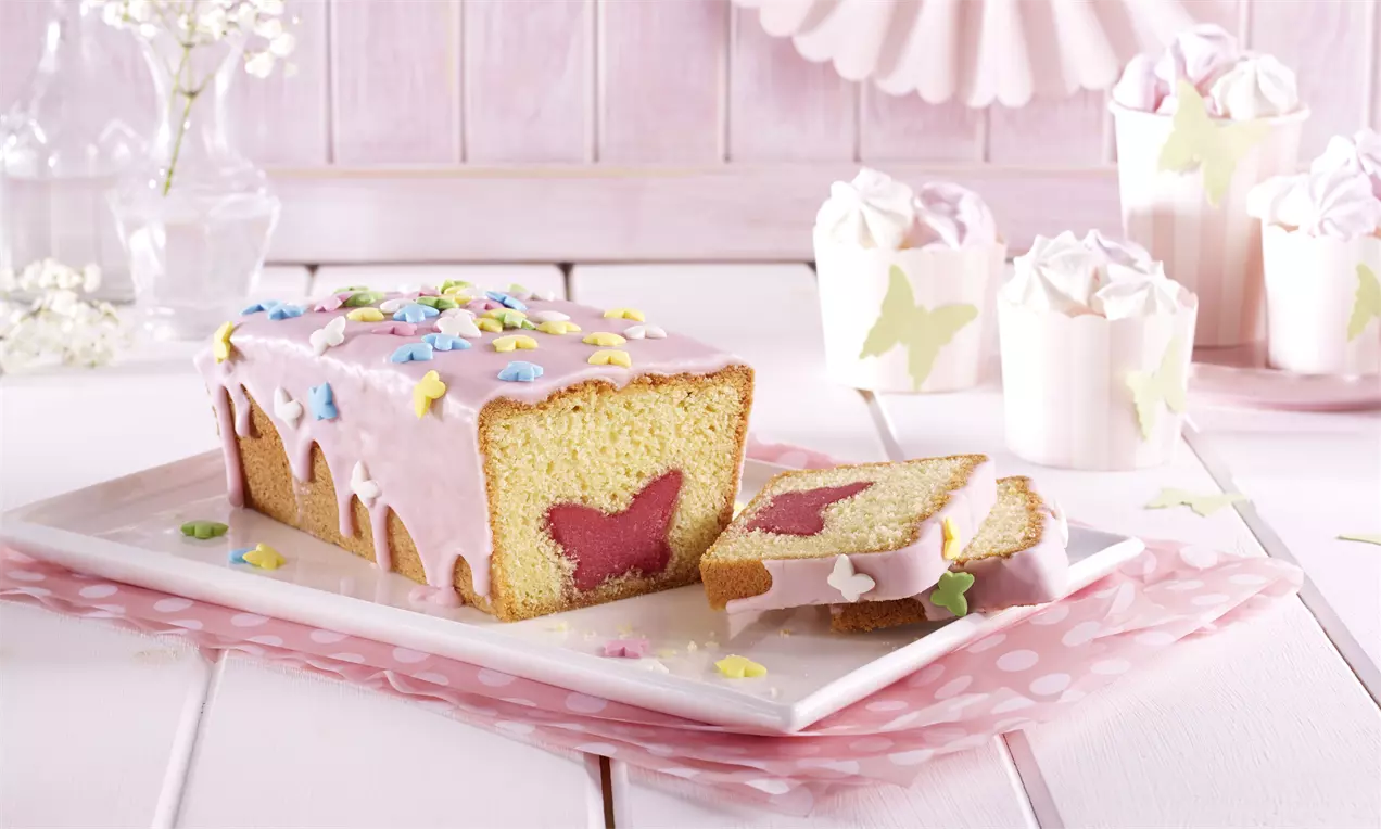 Heller Kastenkuchen mit rosa Zuckerglasur, bunten Streuseln und rosa Schmetterlingsmotiv im Anschnitt auf weißer Platte mit rosa Stoffunterlage